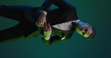 vertical video with colorful lights of sexy young groom with hands in pockets, adjusting tuxedo, crossing arms and looking to side and down