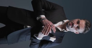 elegant young man in spotlight wearing his bowtie open, rubbing palms, adjusting sleeves and looking to side while fixing tuxedo