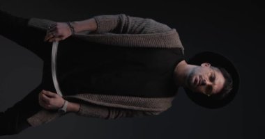 vertical video with flash of cool bearded casual man with glasses and hat rubbing palms, adjusting sweater, looking to side on dark background