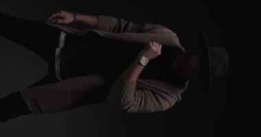 reel of sexy casual man with glasses and hat, adjusting sweater, folding arms and making silence gesture, looking down on dark background