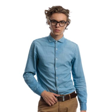 Portrait of sexy casual man pulling his belt with tough attitude, standing, wearing eyeglasses against white studio background