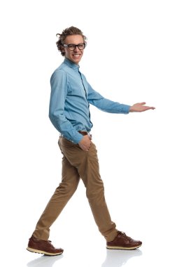 Full body picture of handsome casual man walking with hand in pocket while pointing, standing, wearing eyeglasses against white studio background