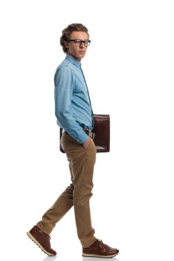 Full body picture of attractive casual man holding briefcase while walking with hand in pocket, standing, wearing eyeglasses against white studio background