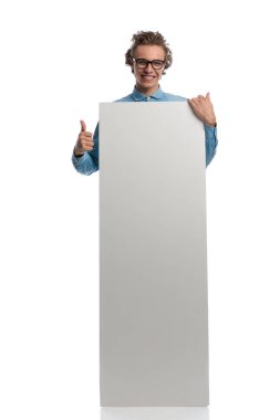 Full body picture of young casual man presenting billboard while giving a thumbs up, standing, wearing eyeglasses against white studio background