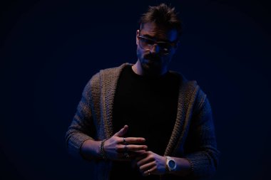 mysterious bearded man with sunglasses looking down and pulling finger while confidently posing on dark background