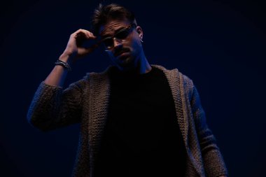 portrait of attractive unshaved man wearing wool cardigan and adjusting sunglasses on dark background
