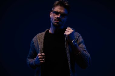 sexy young guy with cool hairstyle pulling and adjusting cardigan while posing on dark background, portrait