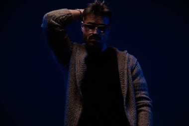 unshaved cool man with cardigan and sunglasses holding hand behind neck and posing on dark background