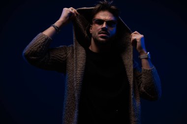portrait of sexy bearded man with sunglasses pulling and adjusting hoodie on dark background 