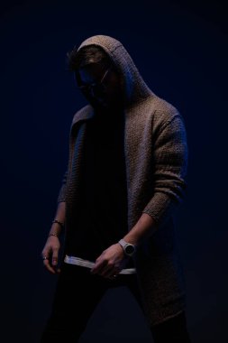 stylish young guy with sunglasses wearing hoodie and looking down while posing in a fashion way on dark background 