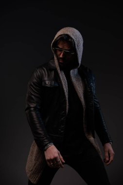 sexy bearded man with glasses looking to side and confidently posing while wearing leather jacket and wool cardigan with hood on grey background