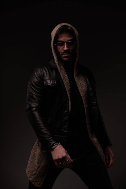 confident bearded man with glasses wearing leather jacket and wool cardigan and posing in a cool manner on grey background