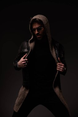 mysterious cool guy adjusting and pulling leather jacket and wool cardigan while posing in a fashion way on grey background