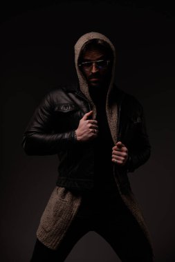 mysterious cool man with glasses pulling and adjusting leather jacket on grey background