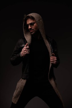 stylish confident man with glasses arranging leather jacket and looking to side on grey background