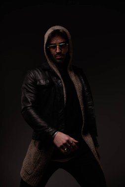 mysterious young man with glasses wearing wool cardigan and leather jacket and posing in a confident way on grey background