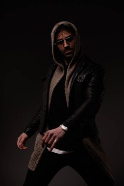 cool young man with glasses posing in a confident manner while wearing leather jacket and wool cardigan with hoodie on grey background