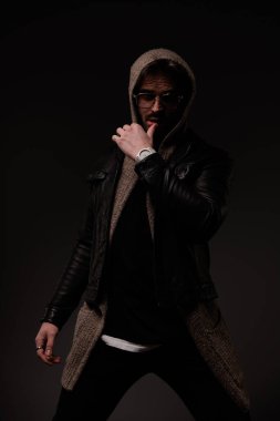 sexy unshaved guy sensually touching lips and posing while wearing leather jacket and wool cardigan with hoodie on grey background