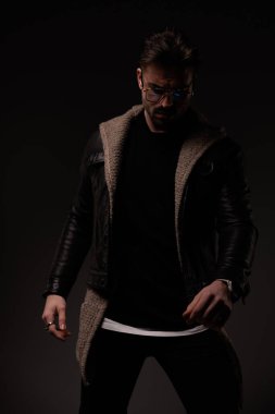 bearded cool guy with stylish hairstyle posing while looking down and wearing leather jacket and wool cardigan on grey background