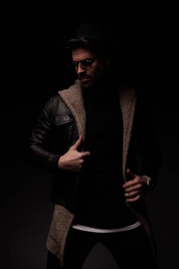 mysterious cool guy looking away and adjusting leather jacket and wool cardigan on grey background