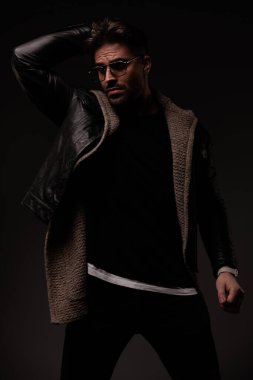 sexy young man with glasses holding arm behind head in a cool way and posing while looking to side on grey background