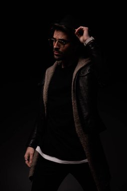dramatic cool man with glasses looking to side and arranging hat on grey background