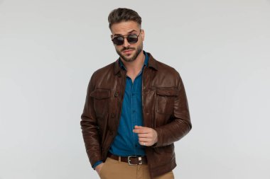 portrait of a attractive casual man posing with macho attitude and hand in pocket, standing, wearing sunglasses against gray studio background