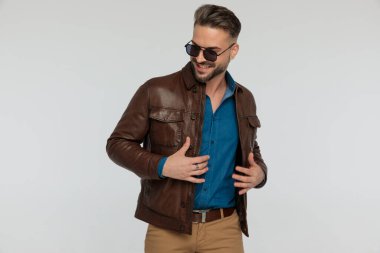 portrait of a young casual man feeling happy and fixing his jacket, standing, wearing sunglasses against gray studio background