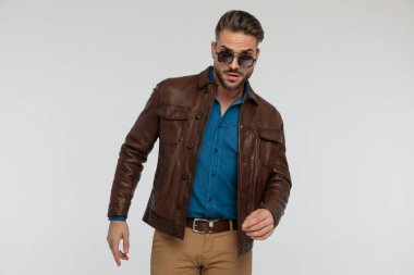 portrait of a sexy casual man posing with tough vibe , standing, wearing sunglasses against gray studio background