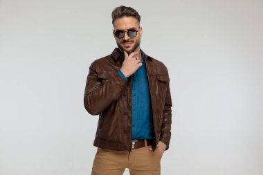 portrait of a young casual man scratching his chin and putting one hand in pocket, standing, wearing sunglasses against gray studio background