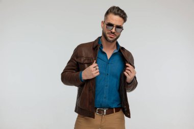 portrait of a handsome casual man adjusting his jacket with tough style, standing, wearing sunglasses against gray studio background