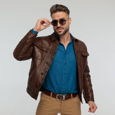 portrait of a young casual man adjusting his sunglasses and posing, standing, against gray studio background