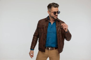 portrait of a handsome casual man looking to side with macho style, standing, wearing sunglasses against gray studio background
