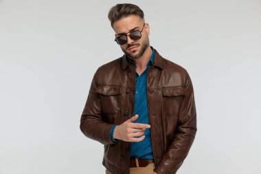 portrait of a young casual man sticking one hand in pocket and posing with style, standing, wearing sunglasses against gray studio background