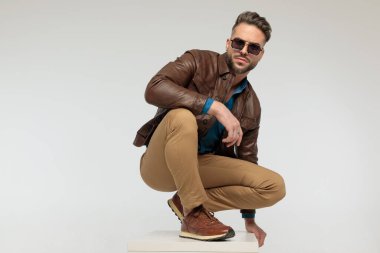 portrait of a sexy casual man resting arms, squatting and looking at the camera, standing, wearing sunglasses against gray studio background