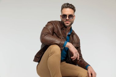 portrait of a young casual man resting arms, squatting with tough attitude, standing, wearing sunglasses against gray studio background