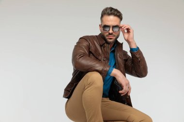 portrait of a handsome casual man resting arms, squatting and fixing sunglasses, standing, against gray studio background