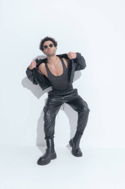 curly young arab man with sunglasses adjusting and pulling leather jacket while laying on a wall on grey background