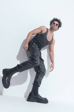 cool fit man wearing leathe rpants and undershirt, laying on a wall and holding one leg up while posing in a side view pose