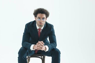 portrait of handsome businessman leaning forward and crossing fingers, sitting, wearing eyeglasses against gray studio background