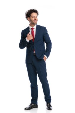 elegant young businessman in navy blue suit looking to side, smiling, fixing suit and holding hand in pocket on white background in studio
