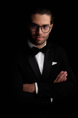 happy elegant groom with glasses in black tuxedo folding arms and smiling in front of white background in studio