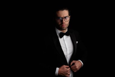 sexy young best man with eyeglasses adjusting and closing black tuxedo in front of black background in studio