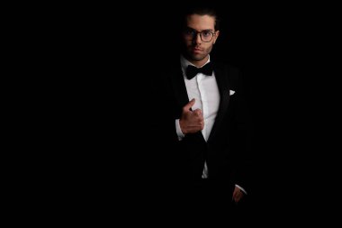 portrait of sexy handsome man with glasses pulling and adjusting black tuxedo in a confident way on black background in studio