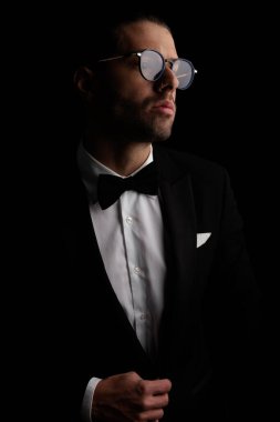 handsome elegant man with glasses looking to side while fixing tuxedo in front of black background in studio