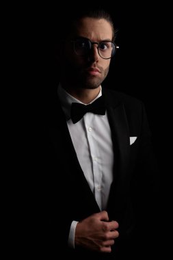 elegant best man with glasses adjusting and buttoning black tuxedo in front of black background