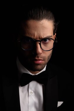 serious elegant groom with glasses in tuxedo frowning in front of black background in studio