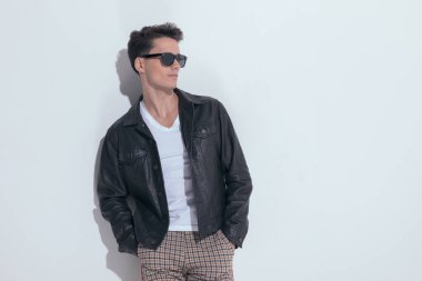 Portrait of  young casual man looking away with hands in pockets, standing, wearing sunglasses in a fashion pose