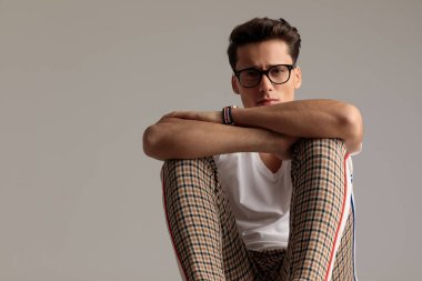 Portrait of young casual man putting hands on his knees, sitting, wearing eyeglasses in a fashion pose