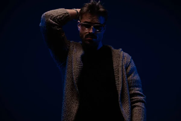 unshaved cool man with cardigan and sunglasses holding hand behind neck and posing on dark background